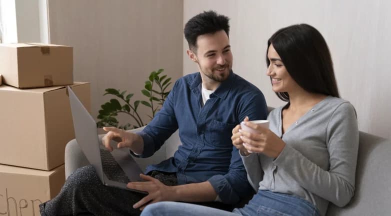 Ein Paar sitzt zwischen Umzugskartons auf dem Sofa und schaut gemeinsam auf einen Laptop