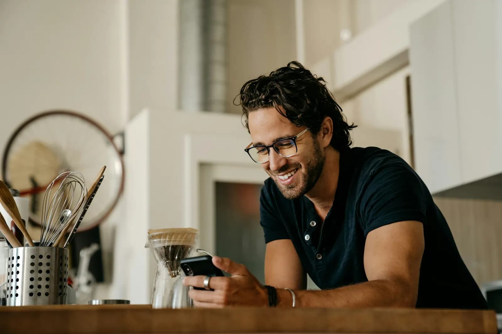 Ein Mann mit Brille steht in seiner Küche und lächelt über eine Nachricht auf seinem Handy